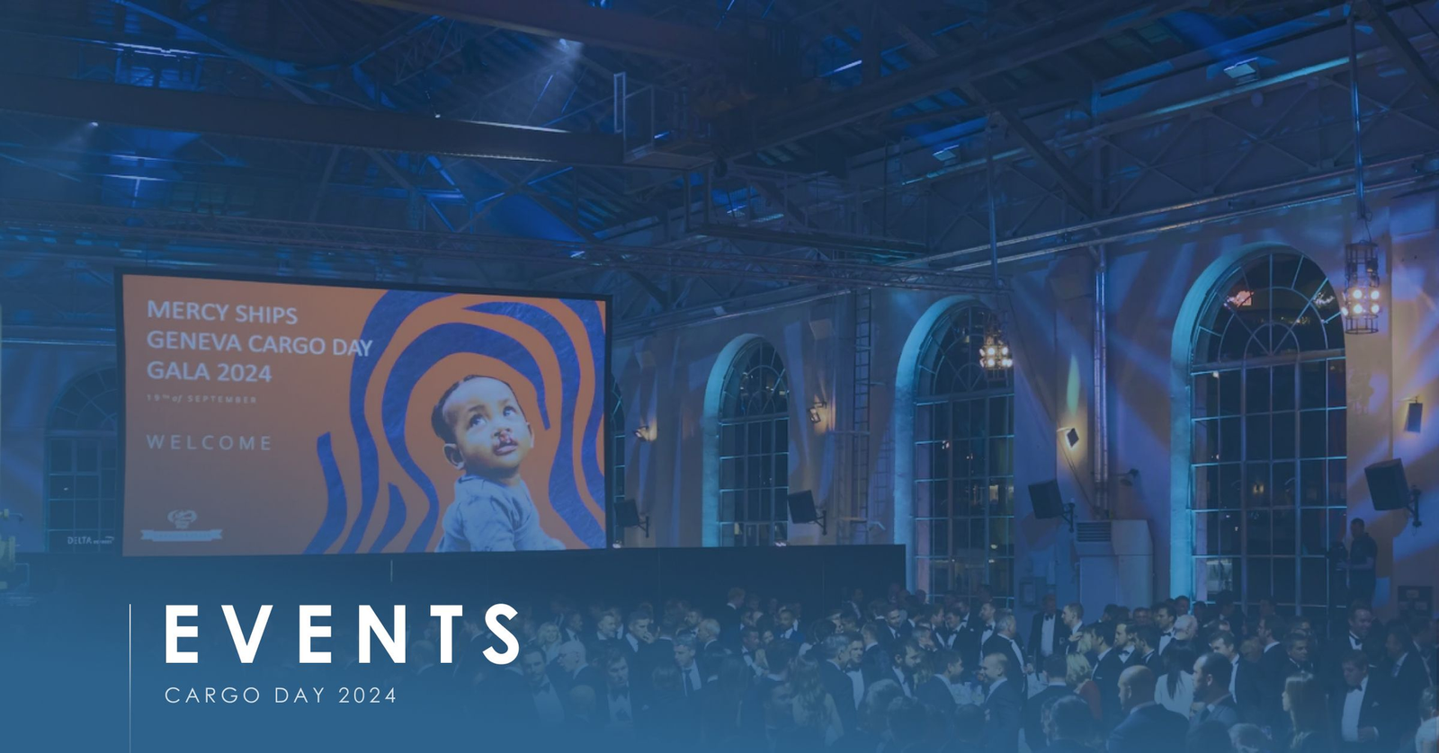 Mercy Ships Geneva Cargo Day Gala 2024 welcome screen above a black-tie crowd in an arched hall